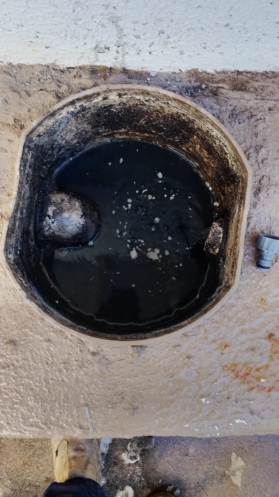 Caixa de gordura redonda aberta, com água escura e um bloco de gordura endurecida e esbranquiçada preso na borda