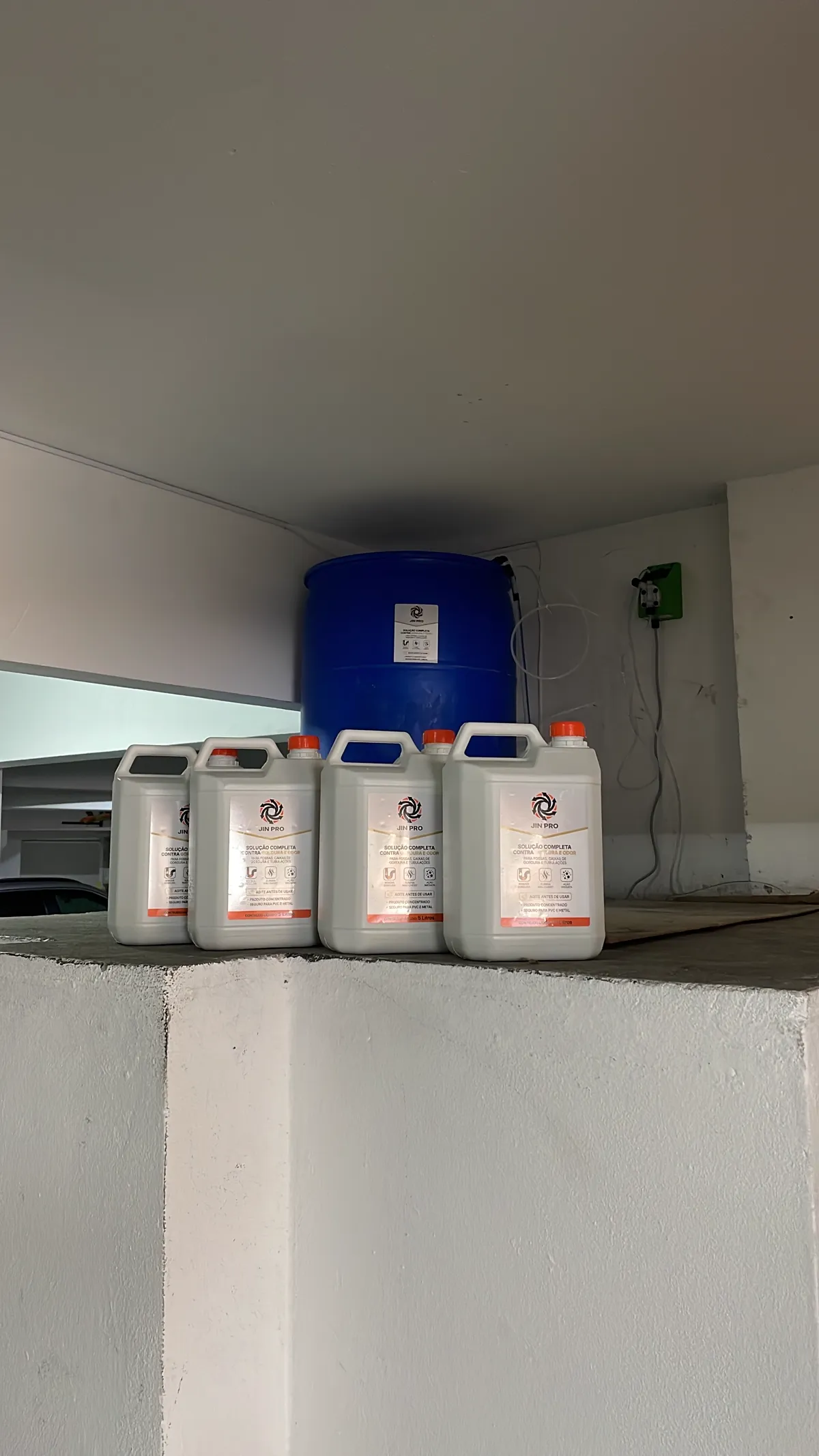 Quatro galões de JIN PRO de 5 litros em frente a um tambor azul com bomba dosadora instalada na parede, o formato de uso recorrente em condomínio e cozinha comercial