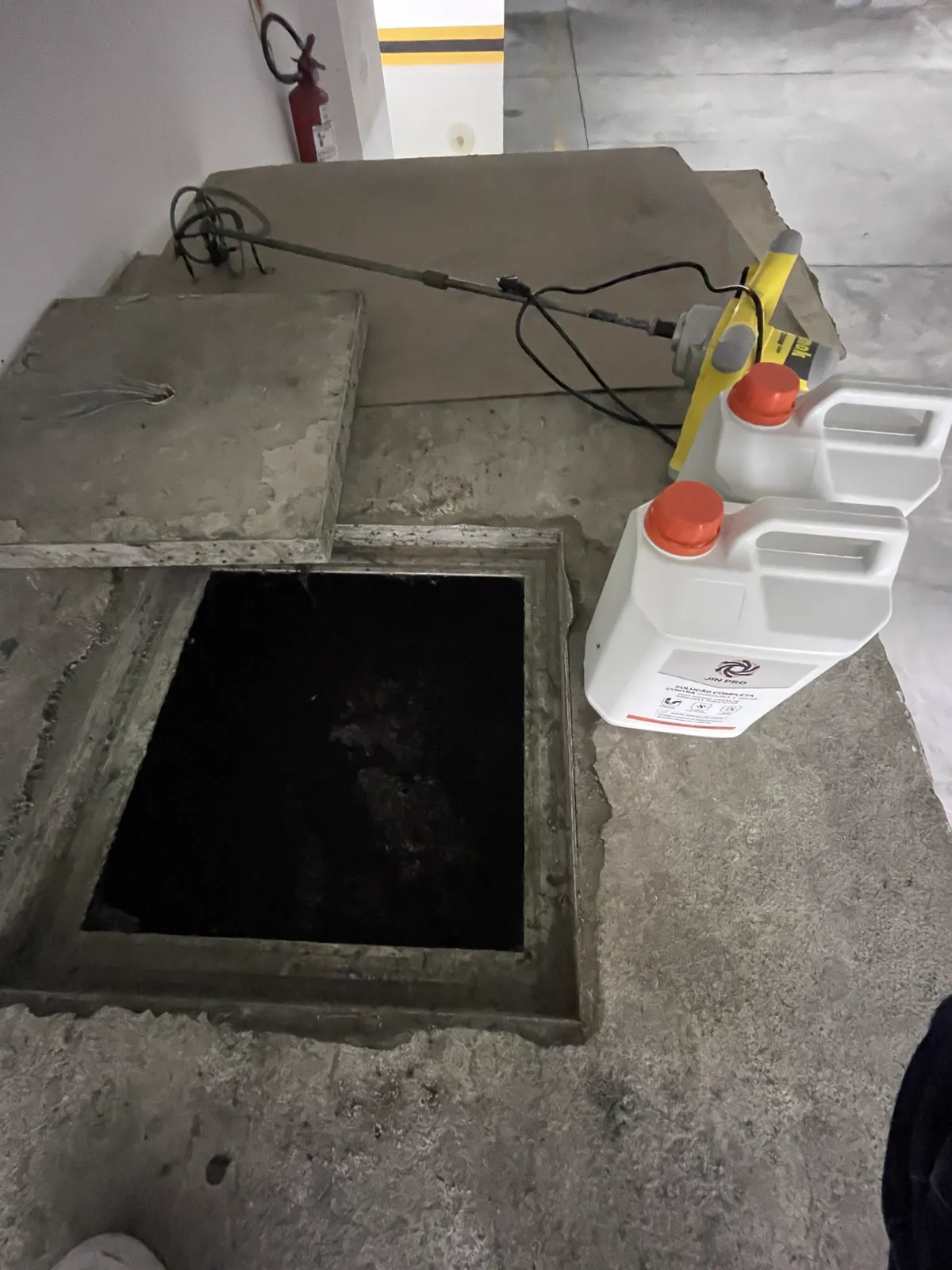 Caixa de gordura aberta no piso de uma garagem, com dois galões de JIN PRO e um misturador elétrico ao lado, prontos para a aplicação